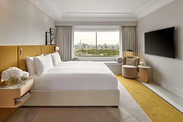 A king bed with white linen faces a large window showing the London skyline and the London Eye, with a wall-mounted television and an armchair beside it.