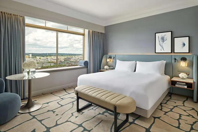 A bedroom with a made king bed, a leather bench at its foot, a round side table and a window looking out over a city skyline.