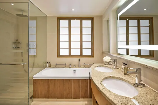 A bathroom with a built-in bathtub beneath a frosted window, a glass-enclosed walk-in shower, and a double vanity with two oval sinks on a granite countertop.