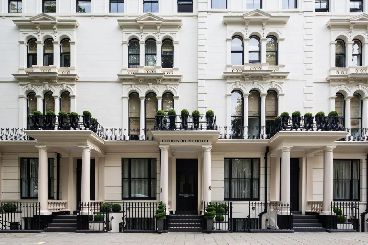Front facade of a white stucco hotel building with tall arched windows, iron balconies with topiary planters, and a sign reading London House Hotel above the entrance.