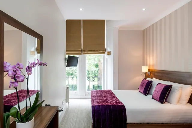 A double bed with purple throws and cushions sits in a room with a tall window looking onto trees and a balcony railing, with a wall-mounted television beside it.