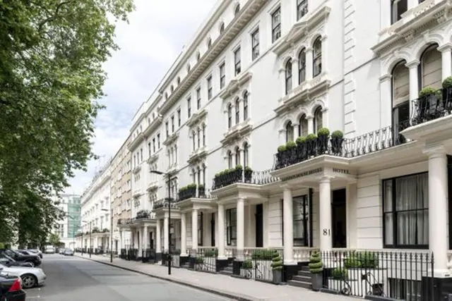 A long white stucco terrace building with iron balconies, planters and tall windows lines a street with parked cars on the left and trees on the far left.