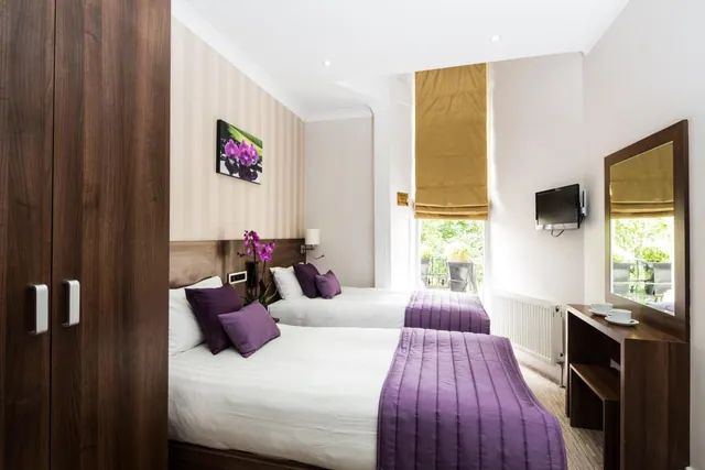 A twin bedroom with two beds dressed in white linen and purple throws, a wall-mounted television, a wooden wardrobe, and a window looking out onto greenery and a balcony with chairs.