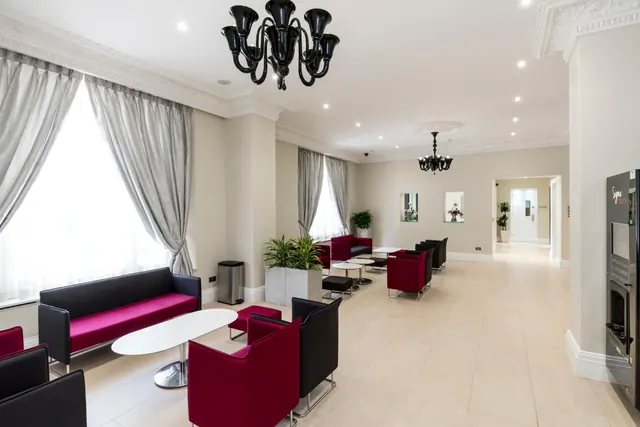 A lounge seating area in a hotel lobby with red and black armchairs, low white tables, black chandeliers and tall windows with grey curtains.