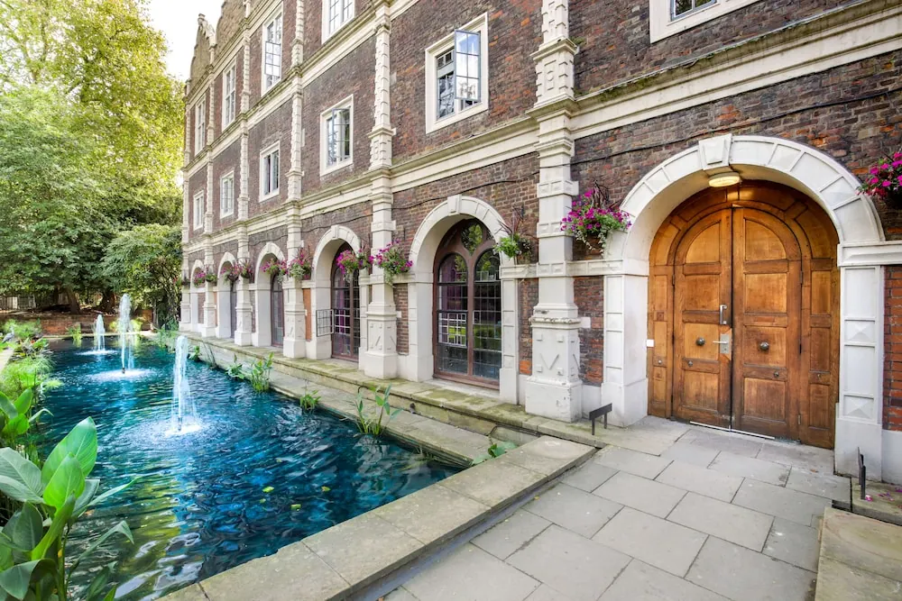 Exterior of a brick building with an arched wooden entrance door, a long water channel with fountains, and flower boxes on the upper windows.