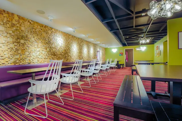 A dining room with purple banquette seating, white chairs and dark wooden tables on a striped carpet, with a textured wooden-block wall and green walls.