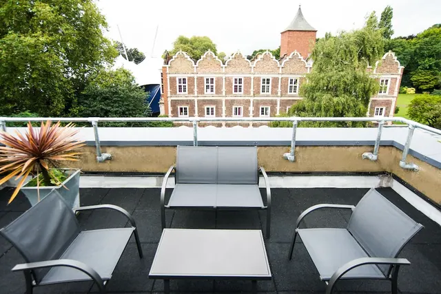 A furnished rooftop terrace with grey metal chairs, a two-seat sofa and a low table, overlooking trees and a historic brick building with gabled facade.