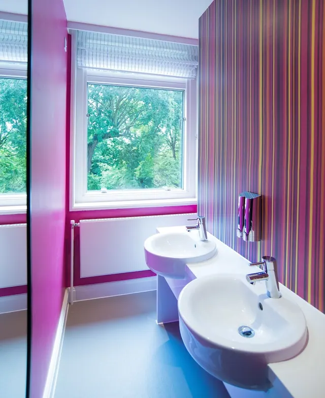 A bathroom with two white basins on a shared counter, striped wallpaper on one wall, and a window showing trees outside.