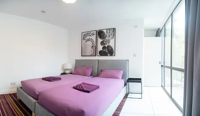 A bedroom with two beds with grey upholstered headboards and purple bedding, black-and-white wall art, and floor-to-ceiling windows on the right showing foliage outside.