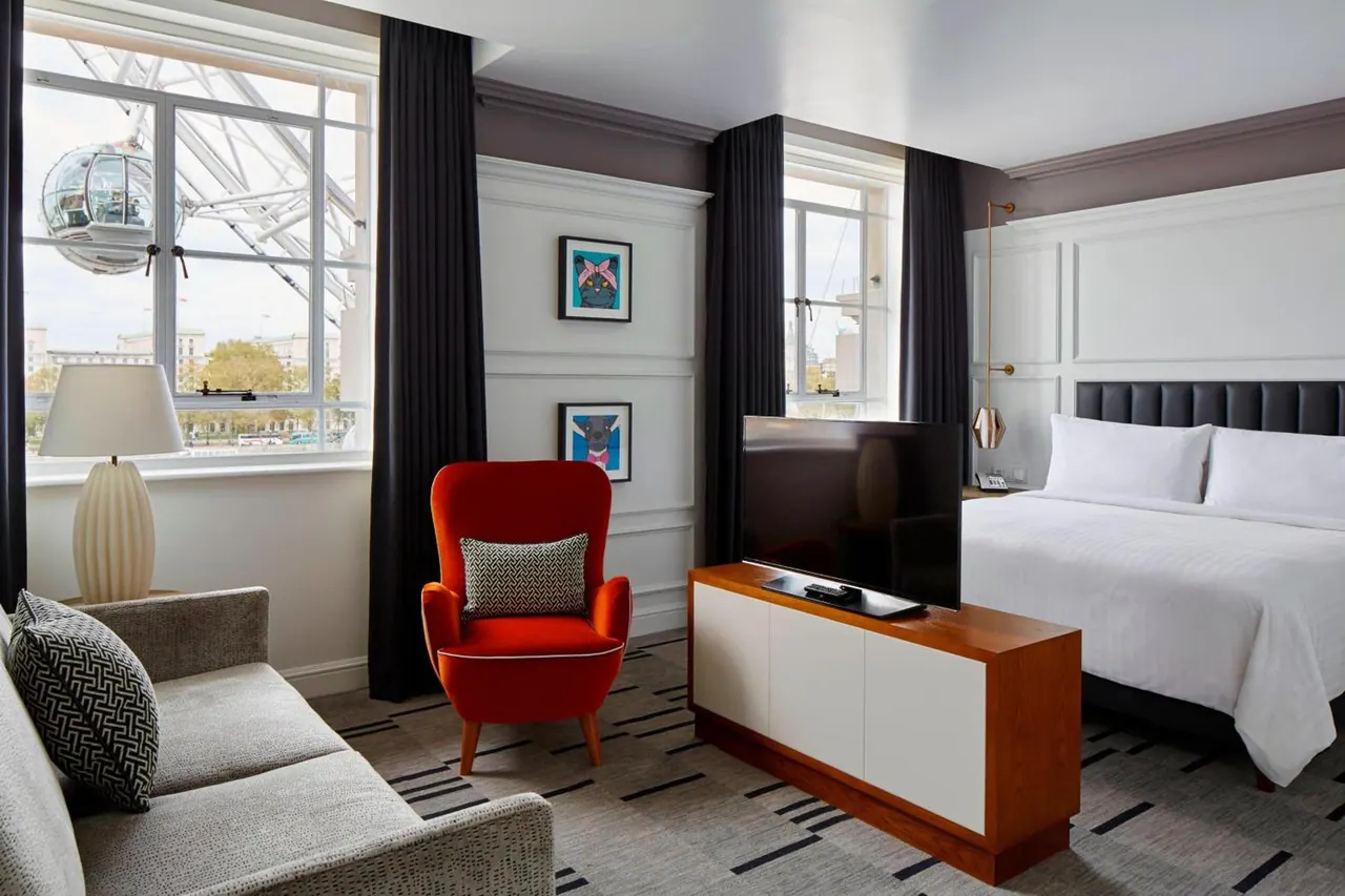 A hotel room with a king bed, a red armchair, a grey sofa and a wall-mounted television, with a window showing the London Eye.