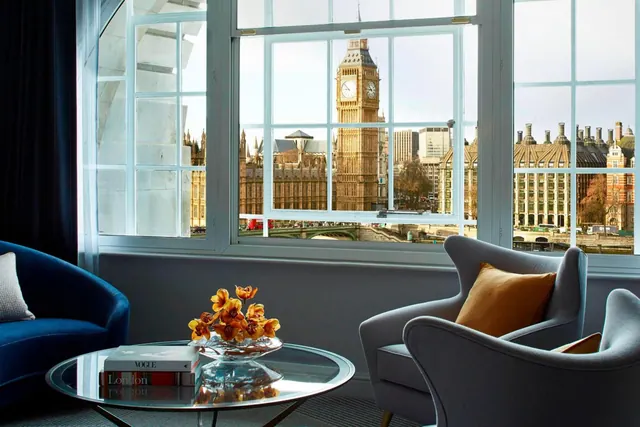 A living area with grey armchairs and a glass coffee table holding books and orange flowers, with windows framing Big Ben and the Palace of Westminster across the river.
