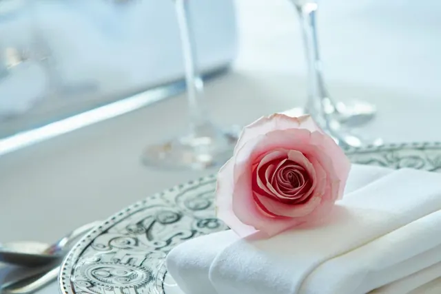 A pink rose rests on a folded white napkin beside a patterned plate, cutlery and wine glasses on a dining table.