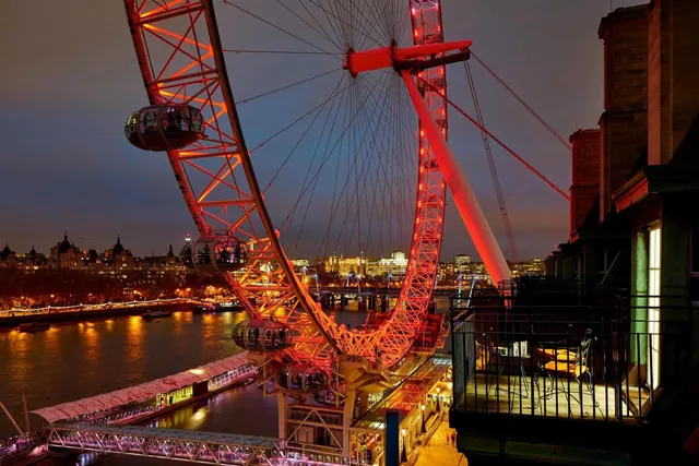 Night view of the illuminated London Eye and the River Thames from a balcony with chairs and a metal railing.
