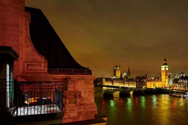 A night view from a stone balcony over the River Thames towards Big Ben and the Palace of Westminster, both lit up after dark.