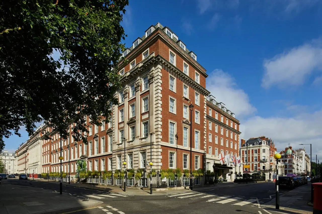 Exterior of a large red-brick hotel building with stone trim and flags at the entrance, viewed across a street with pedestrian crossings on a sunny day.