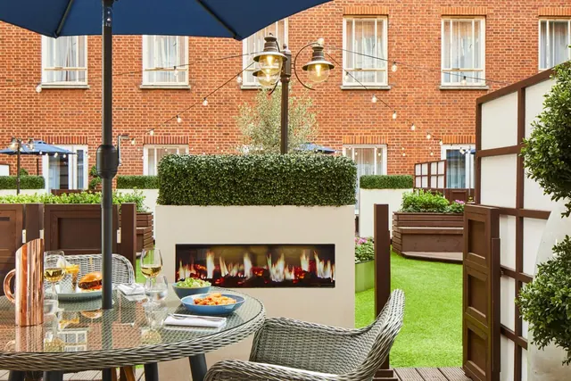 An outdoor dining table with wine glasses, food and a blue parasol stands on a terrace beside a gas fire in a planter, with a brick building behind.