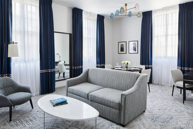 A living area with a grey sofa, grey armchair, white oval coffee table, navy curtains and a small dining table set for two near the windows.