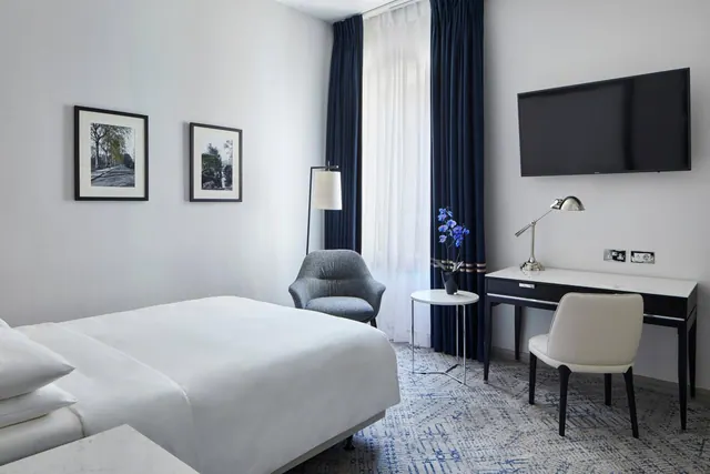 A hotel bedroom with a made white bed, a grey armchair, a wall-mounted television, and a desk with a lamp and chair beside navy curtains.