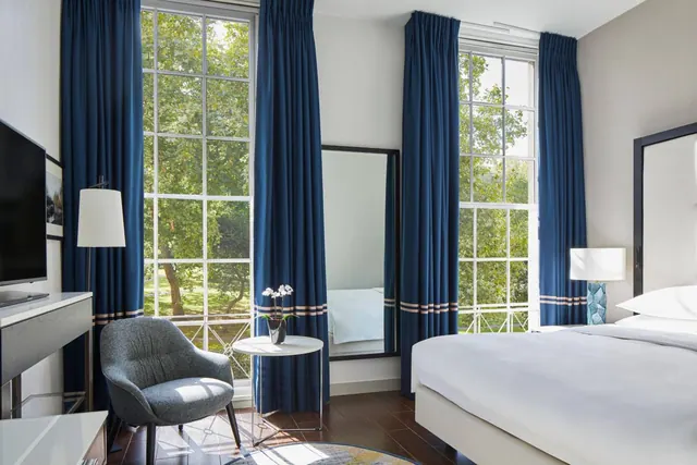 A bedroom with a made king bed, navy curtains, a grey armchair, a round side table with an orchid, and tall windows looking onto trees.