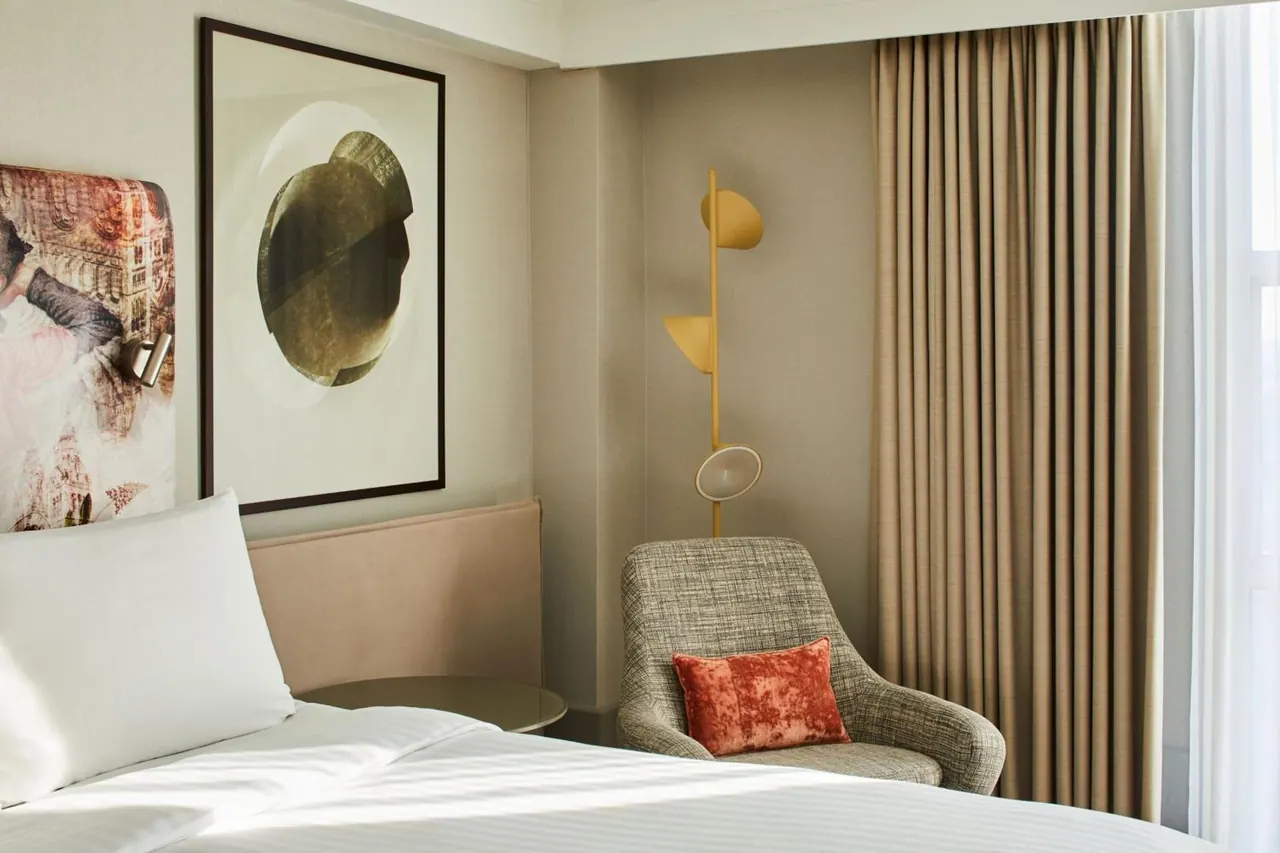 A hotel bedroom with a made bed in the foreground, an upholstered armchair with a red cushion, a gold wall lamp and beige curtains.