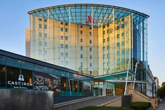 A curved glass-fronted hotel building with a Marriott sign and a Cast Iron Bar & Grill entrance at street level, photographed in early evening light.