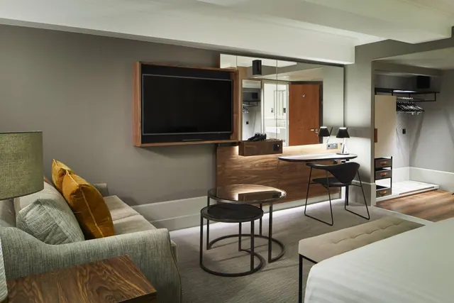 A hotel room seating area with a grey sofa with gold cushions, a wall-mounted television, a desk with a chair, nesting coffee tables, and the foot of a bed at the right.