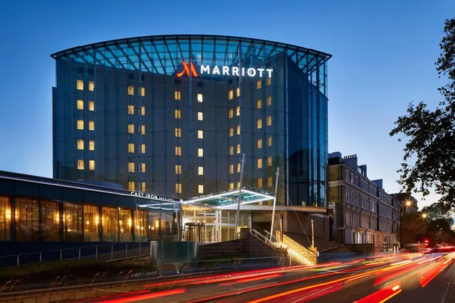 Exterior of a glass-fronted Marriott hotel at dusk with illuminated windows, a Marriott sign on the upper facade, and traffic light trails on the street below.