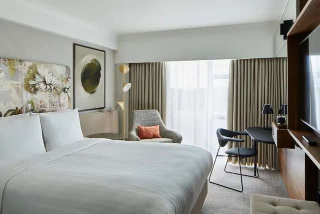 A hotel bedroom with a made double bed, an armchair beside a window with sheer curtains, a desk with a chair, and a wall-mounted television.