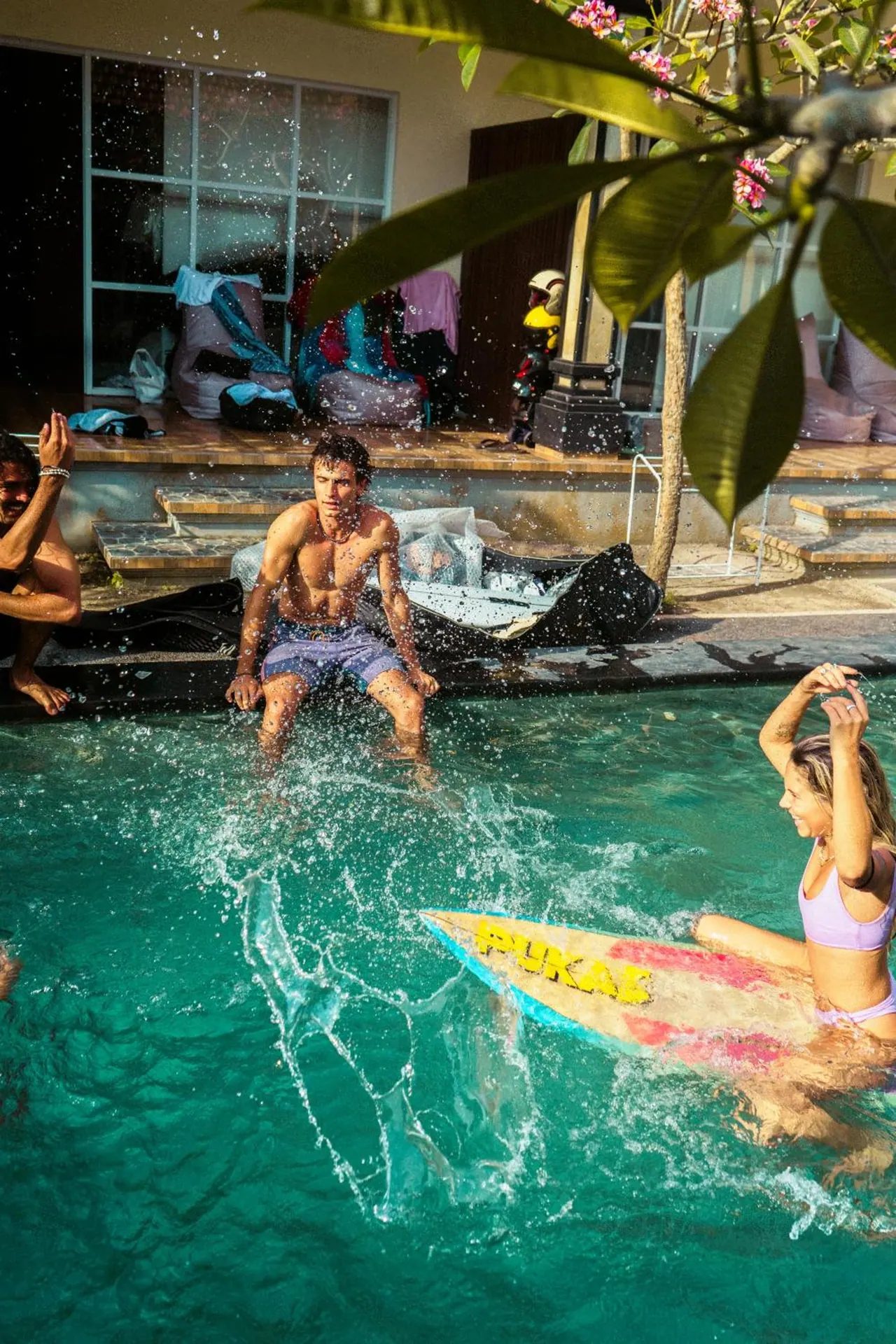 Three people are at a turquoise pool, with a woman sitting on a surfboard in the water and a man seated on the edge while water splashes around them.