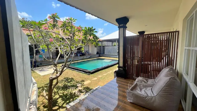 A covered terrace with a grey bean bag chair looks out over a garden with a frangipani tree and a swimming pool.