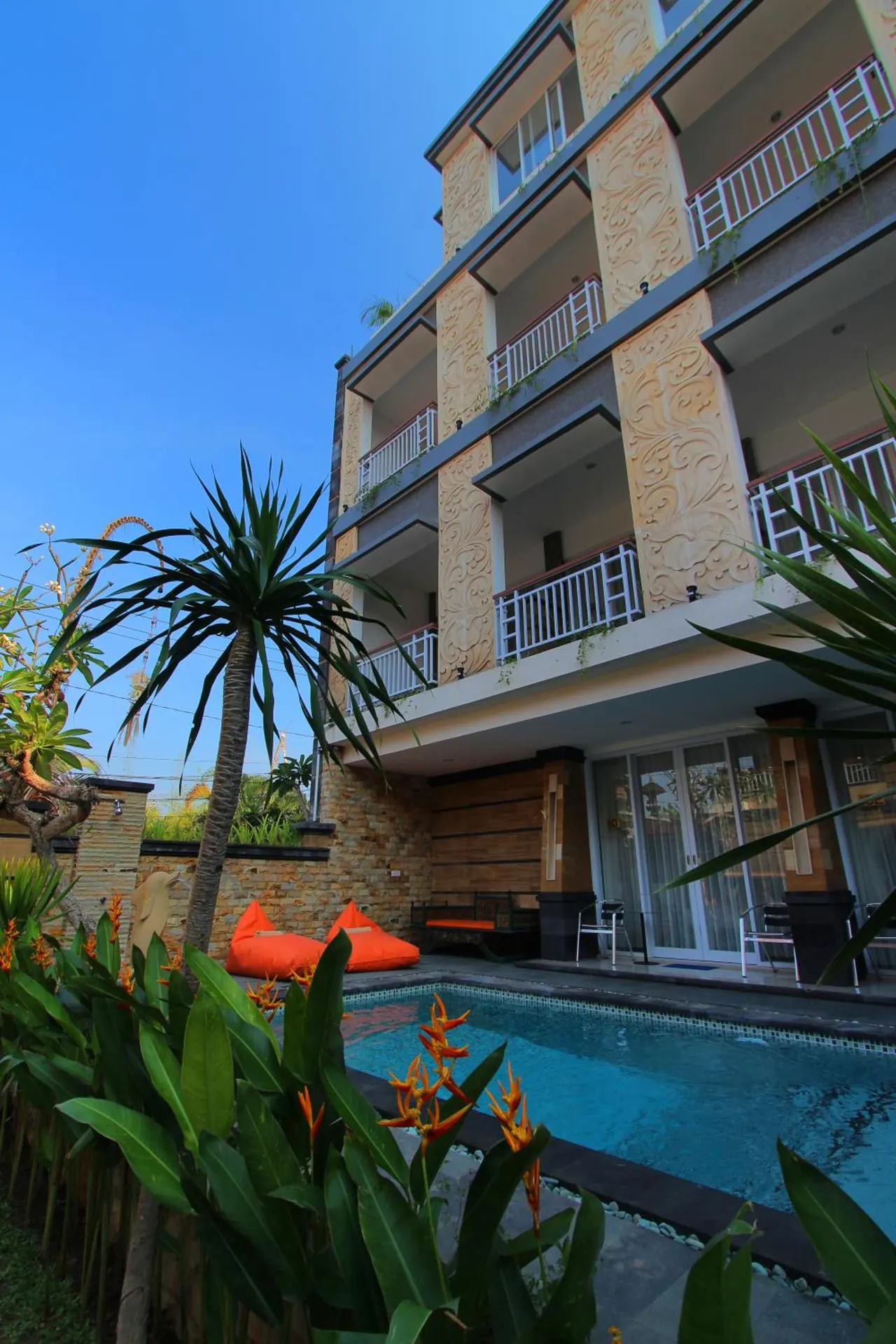 Low-angle view of a four-storey hotel building with carved stone pillars and balconies, with a small swimming pool and orange bean bags in the foreground under a clear blue sky.