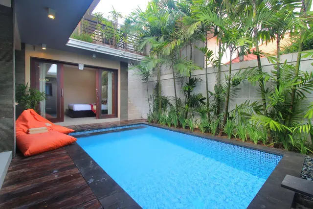 A small private swimming pool on a timber deck with orange bean bags, tall palms along the back wall, and a bedroom visible through sliding glass doors.