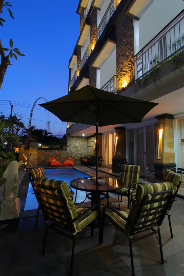 A dusk view of an outdoor terrace with a shaded dining table and striped chairs beside a small pool and a multi-storey building with lit balconies.