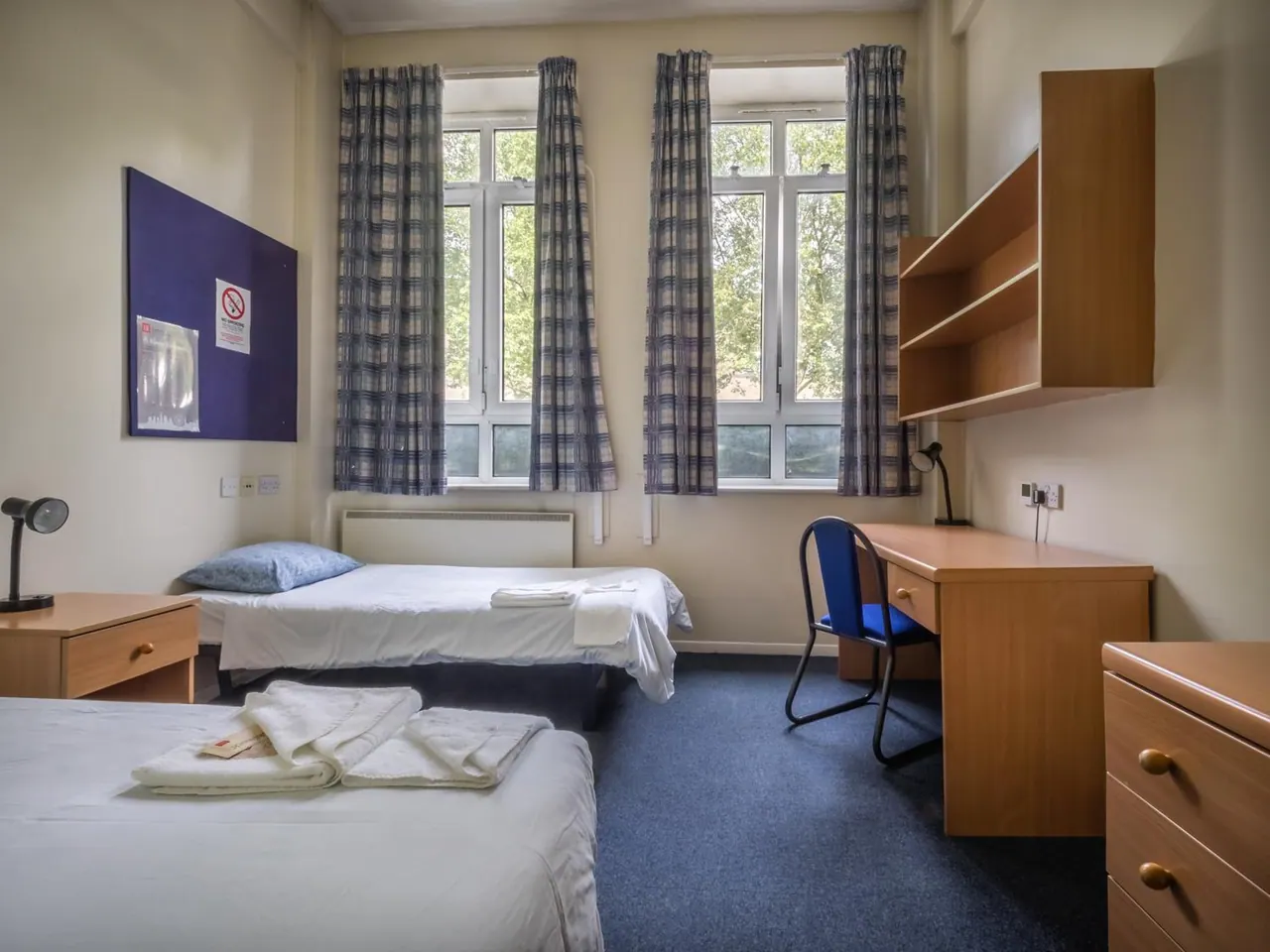 A bedroom with two single beds, a desk and chair, wall shelving, and two curtained windows looking out onto trees.