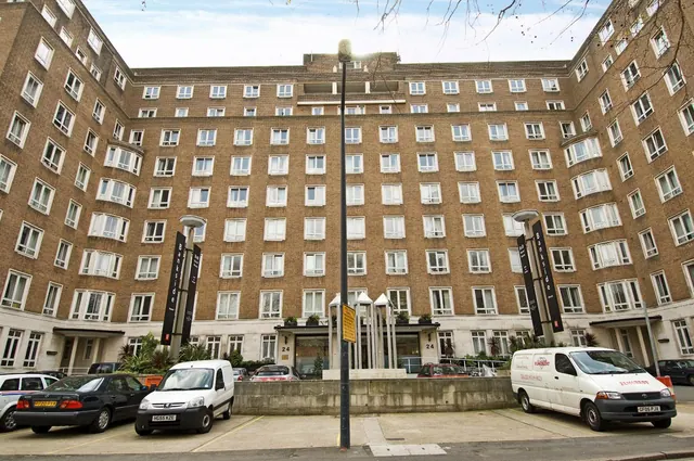 Exterior view of a tall brick building with rows of windows, a street lamp post, a building entrance with planters, and parked cars and a white van in the foreground.