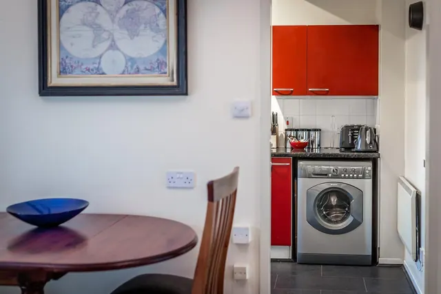 A round wooden dining table and chair sit beside a kitchenette with red cabinets, a washing machine, a kettle and a toaster.
