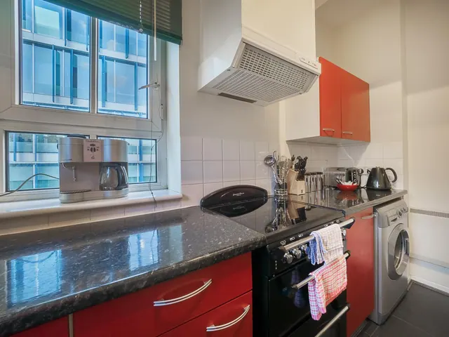 A small kitchen with red cabinets, a black electric hob and oven, a coffee machine and kettle on the worktop, and a window overlooking a modern glass building.