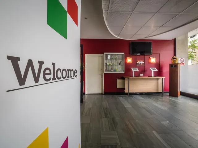 A hotel lobby with a red feature wall, a wall-mounted television, a reception desk with two check-in kiosks, and a large Welcome sign on the left.