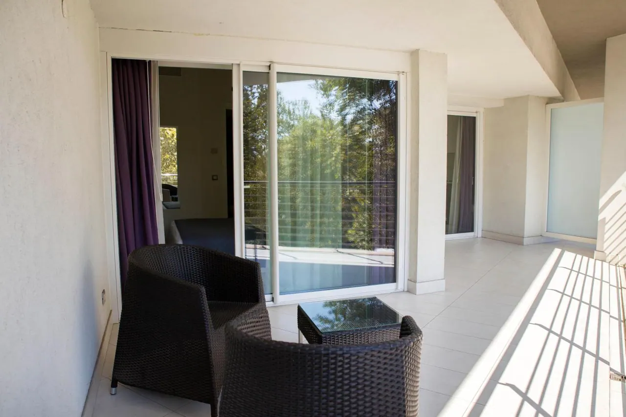 A tiled balcony with two wicker armchairs and a glass side table, with sliding glass doors open to a room and trees visible beyond.