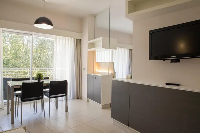 A bright living area with a glass dining table and black chairs, a wall-mounted television, and sliding doors onto a balcony with trees outside.