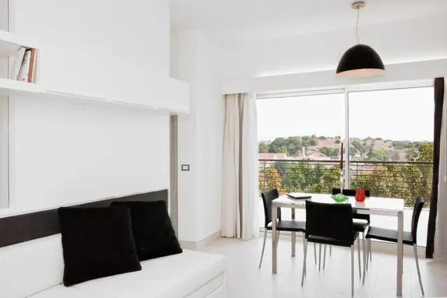 A bright white living area with a white sofa and black cushions, a dining table with black chairs, and a large window looking out onto trees and hills.