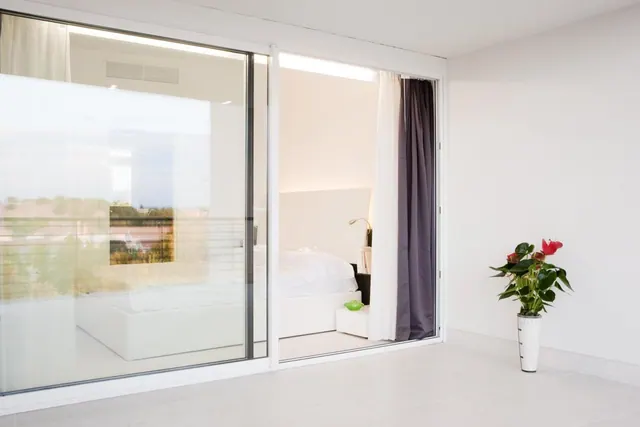 A white bedroom with a double bed is seen through sliding glass doors, with a potted red anthurium plant on the tiled floor in the foreground.