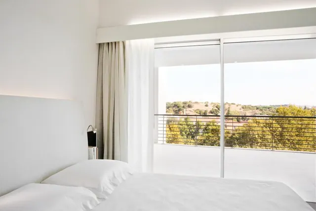 A bedroom with white bedding and a white headboard, with sliding glass doors opening onto a railed balcony and a view of trees and a hillside.