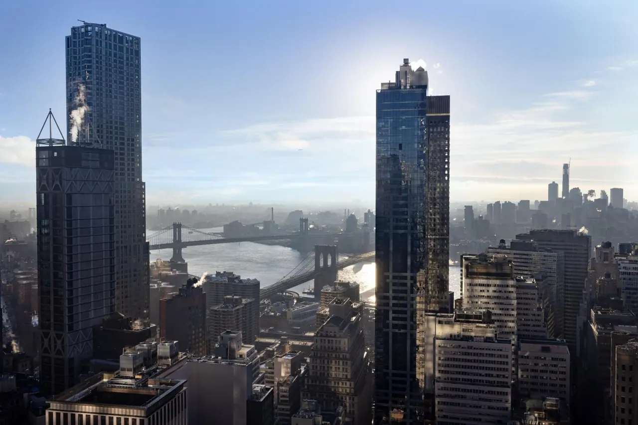 Elevated view over a dense city skyline toward two bridges crossing a river under a hazy blue sky.