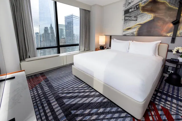 A made-up king bed sits in a hotel room beside a floor-to-ceiling window looking out over city skyscrapers.