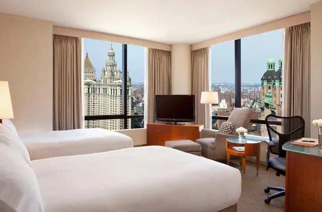 A hotel bedroom with two beds, a television, an armchair and a desk, with windows looking out over a city skyline that includes the Woolworth Building.