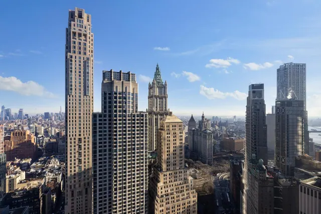 A high-level view over Manhattan towers, including the green-topped Woolworth Building, with a river visible on the right under a clear blue sky.