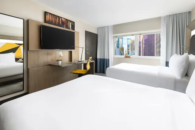 A hotel room with two white beds, a wall-mounted television, a desk with a yellow chair, and a window showing city buildings.