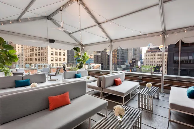 A covered rooftop terrace with grey sofas, coloured cushions, string lights and a view of surrounding city buildings and a Wicked billboard.