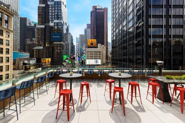 A rooftop terrace with red and navy stools and high tables overlooks Times Square and the surrounding skyscrapers.